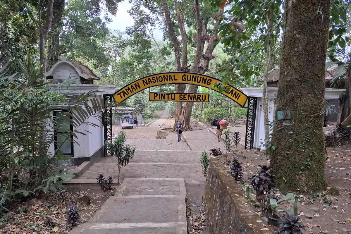 A paved path under a yellow arch reading Taman Nasional Gunung Rinjani, Pintu Senaru, with small white buildings and big trees around it.