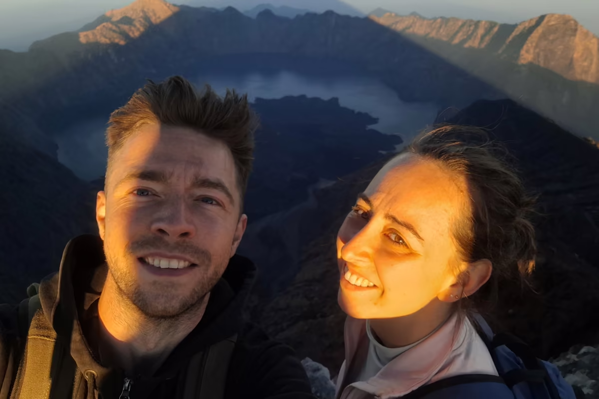 A smiling man and woman taking a selfie in warm early-morning light on the summit, with the crater lake and the caldera rim behind them.