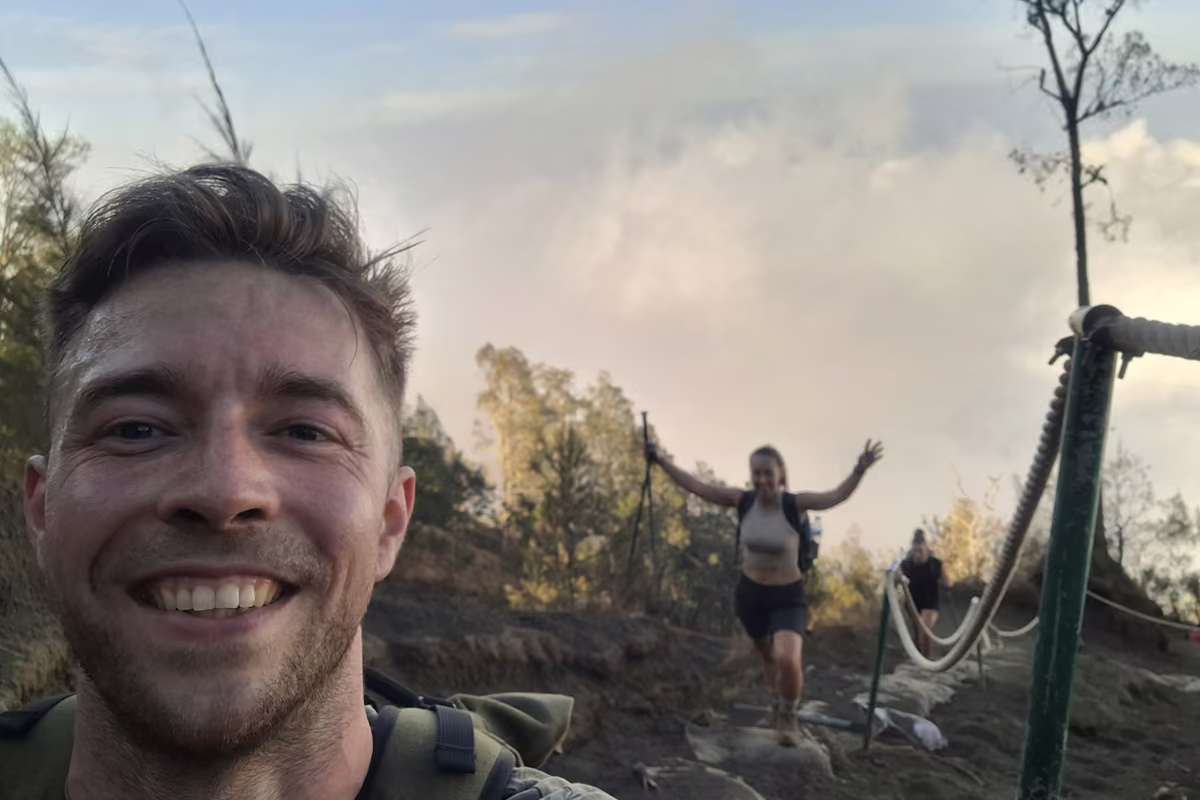 A smiling hiker taking a selfie on a dusty trail, while a fellow hiker behind him raises her arms beside a rope railing and the clouds below.