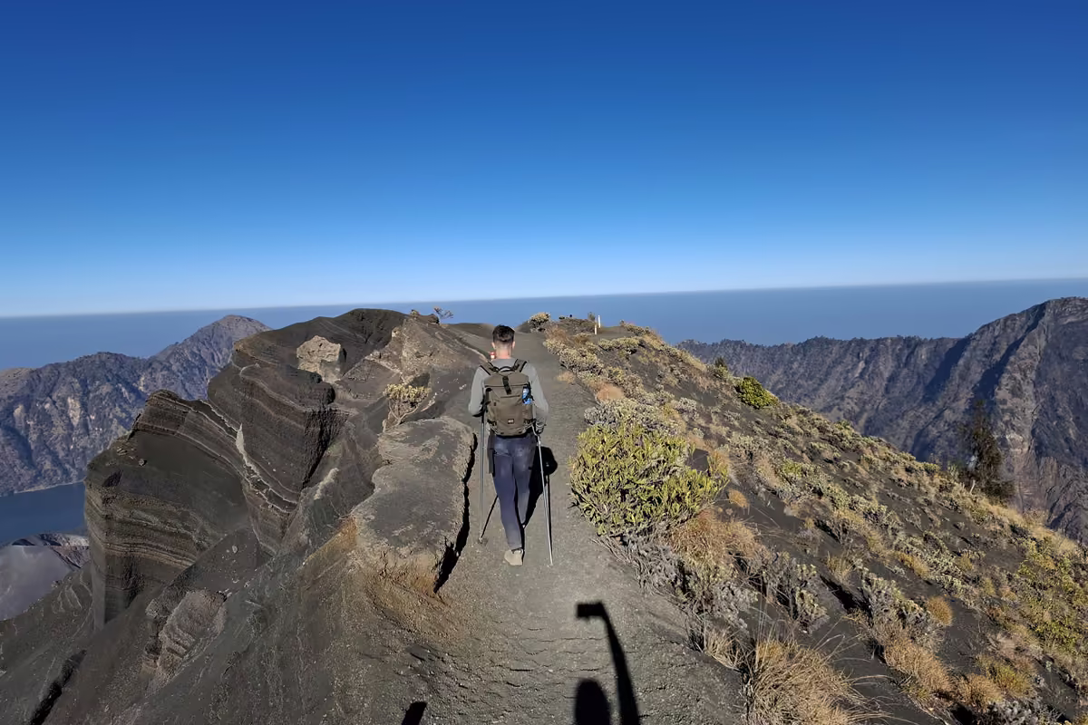 A hiker with trekking poles walking on a path beside layered dark volcanic rock at the top of the mountain, with the caldera ridges below and the sea on the horizon under a deep blue sky.
