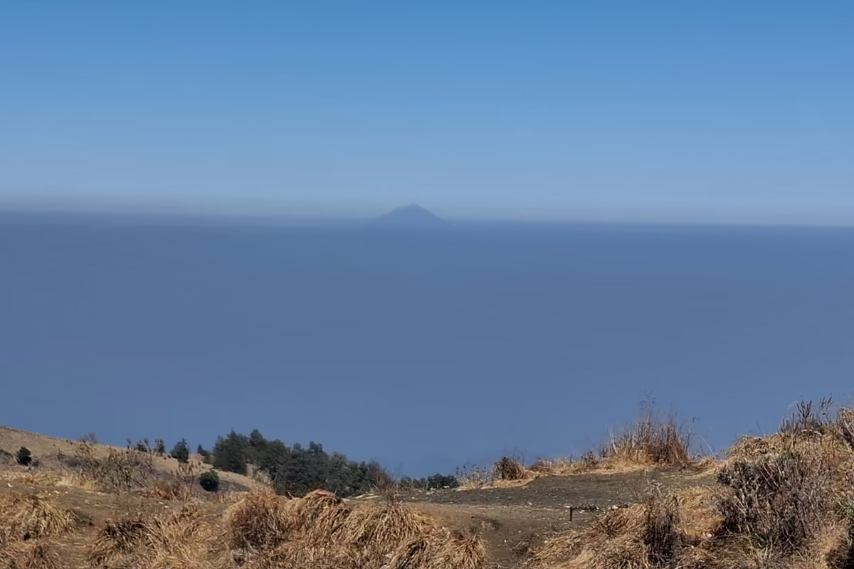 The cone of Mount Agung on Bali rising above a hazy horizon across the sea, seen over dry grass on the Senaru crater rim.