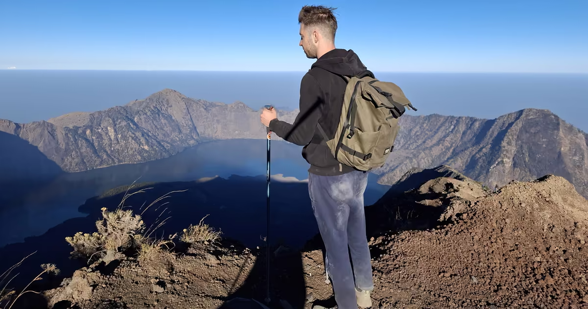 A hiker with a backpack and trekking poles standing on the Mount Rinjani summit, looking out over the caldera, the Segara Anak crater lake and the sea on the horizon.